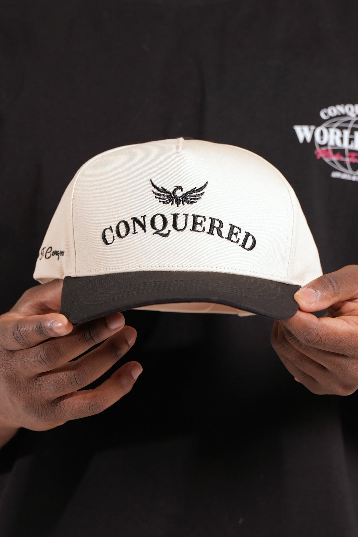 Person holding a cream and black satin lined cap with 'Conquered' text and image logo stitched on against a black shirt background