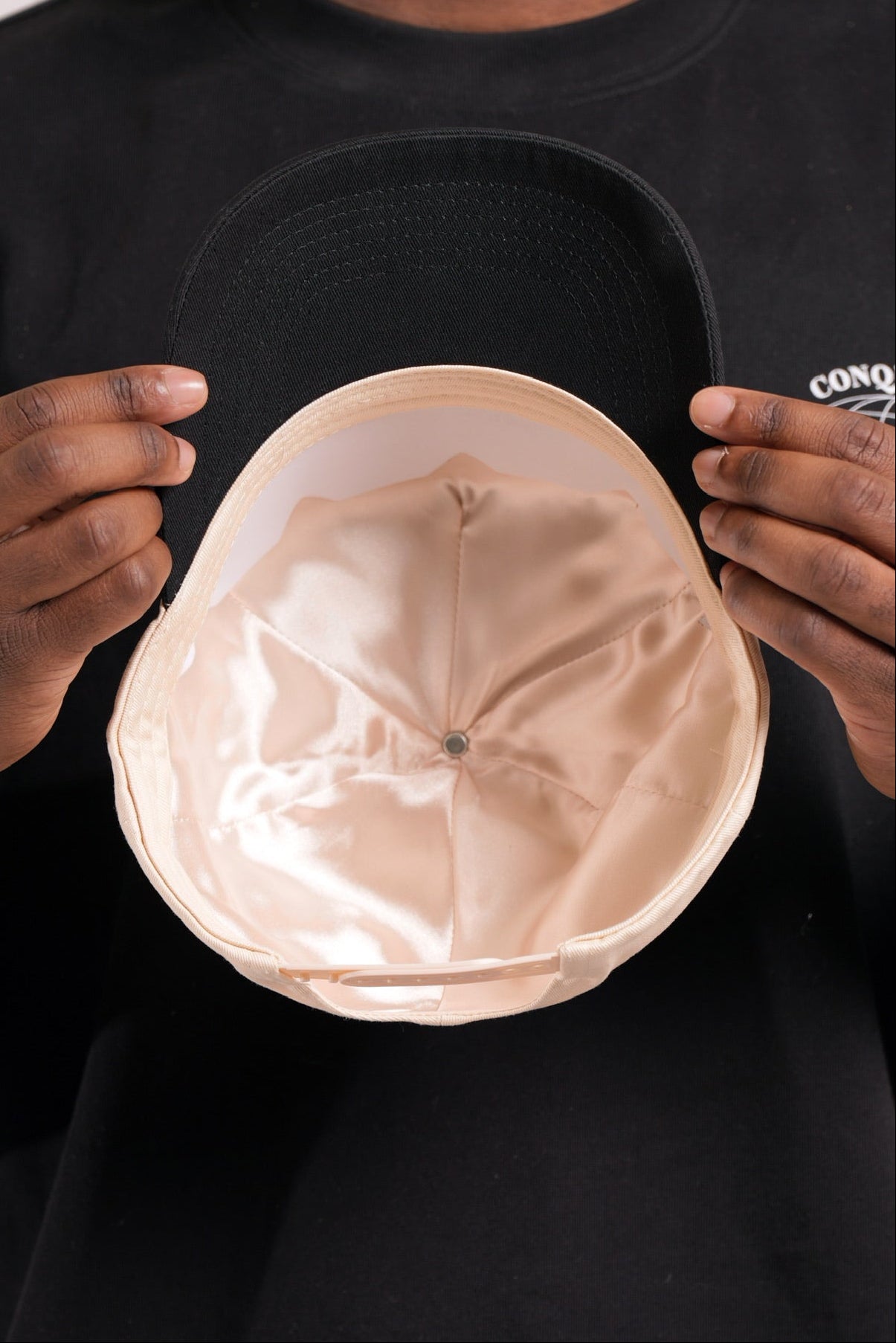 Person holding a cream and black satin lined cap with 'Conquered' text and image logo stitched on against a black shirt background