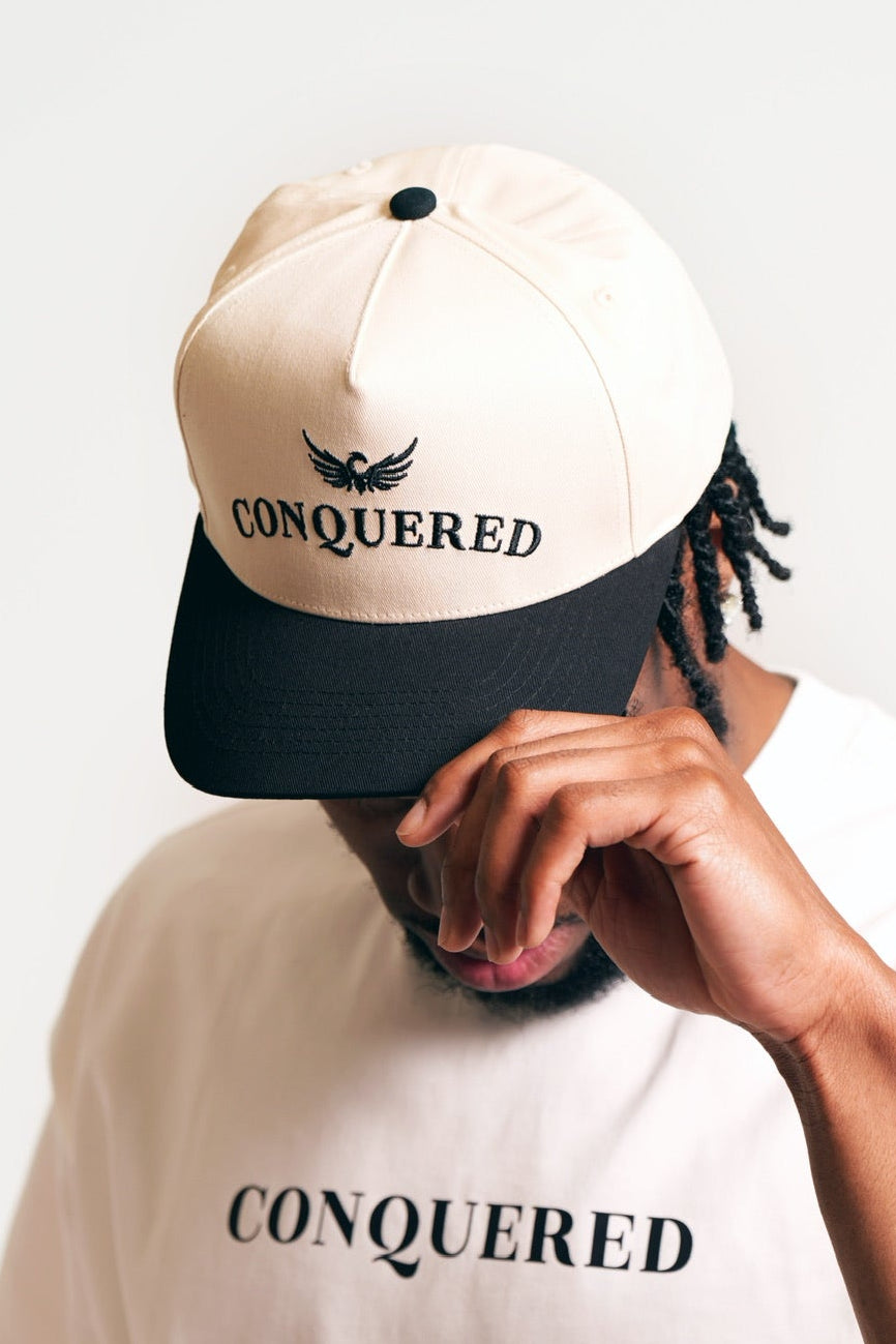 Person wearing a cream and black coloured satin lined cap with 'CONQUERED' word and image branding on a plain white background