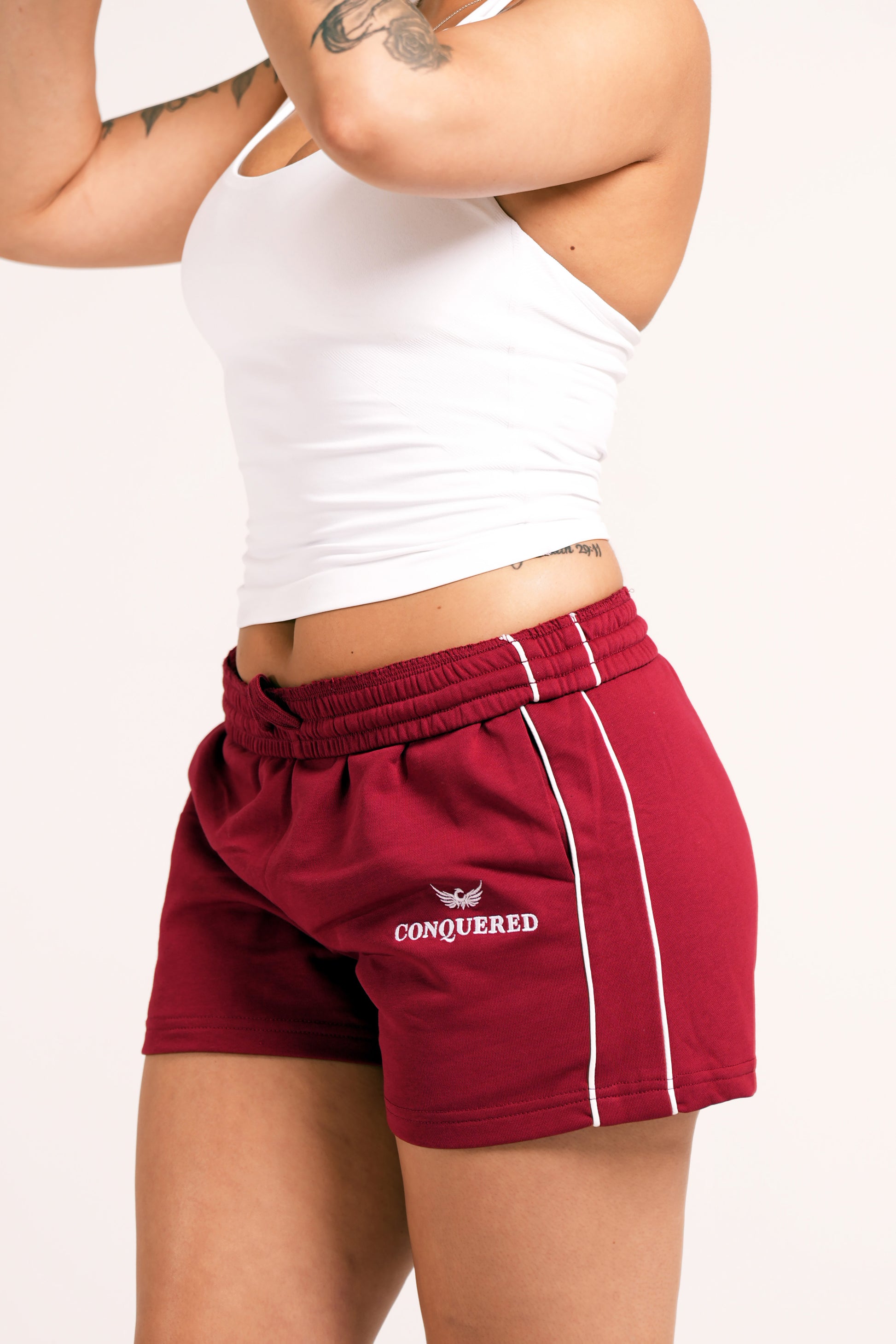 Person wearing crimson red varsity style shorts with white lines on the side with 'Conquered' text and image logo embroidered below the left pocket pairing with a white halter top in front of a plain white background