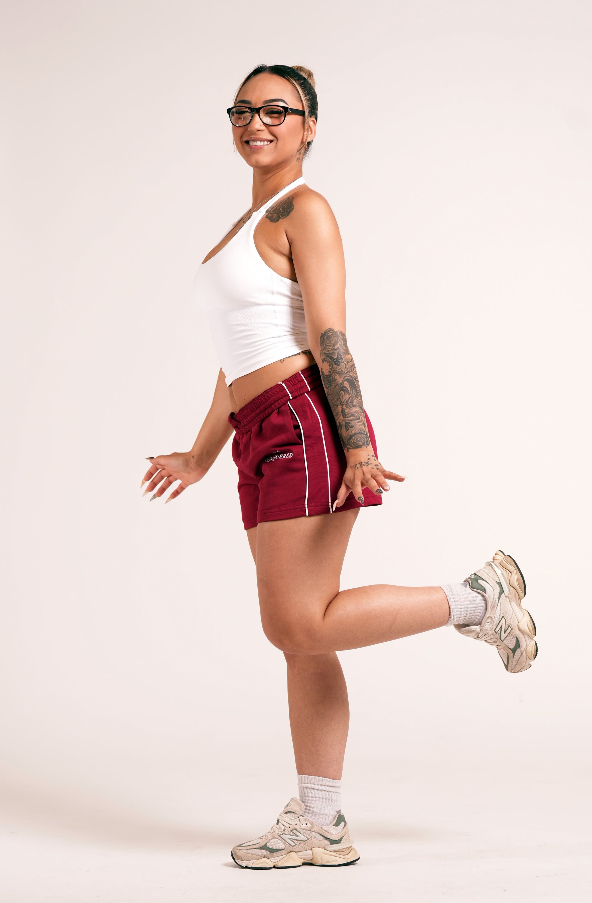 Person wearing crimson red varsity style shorts with white lines on the side with 'Conquered' text and image logo embroidered below the left pocket pairing with a white halter top in front of a plain white background
