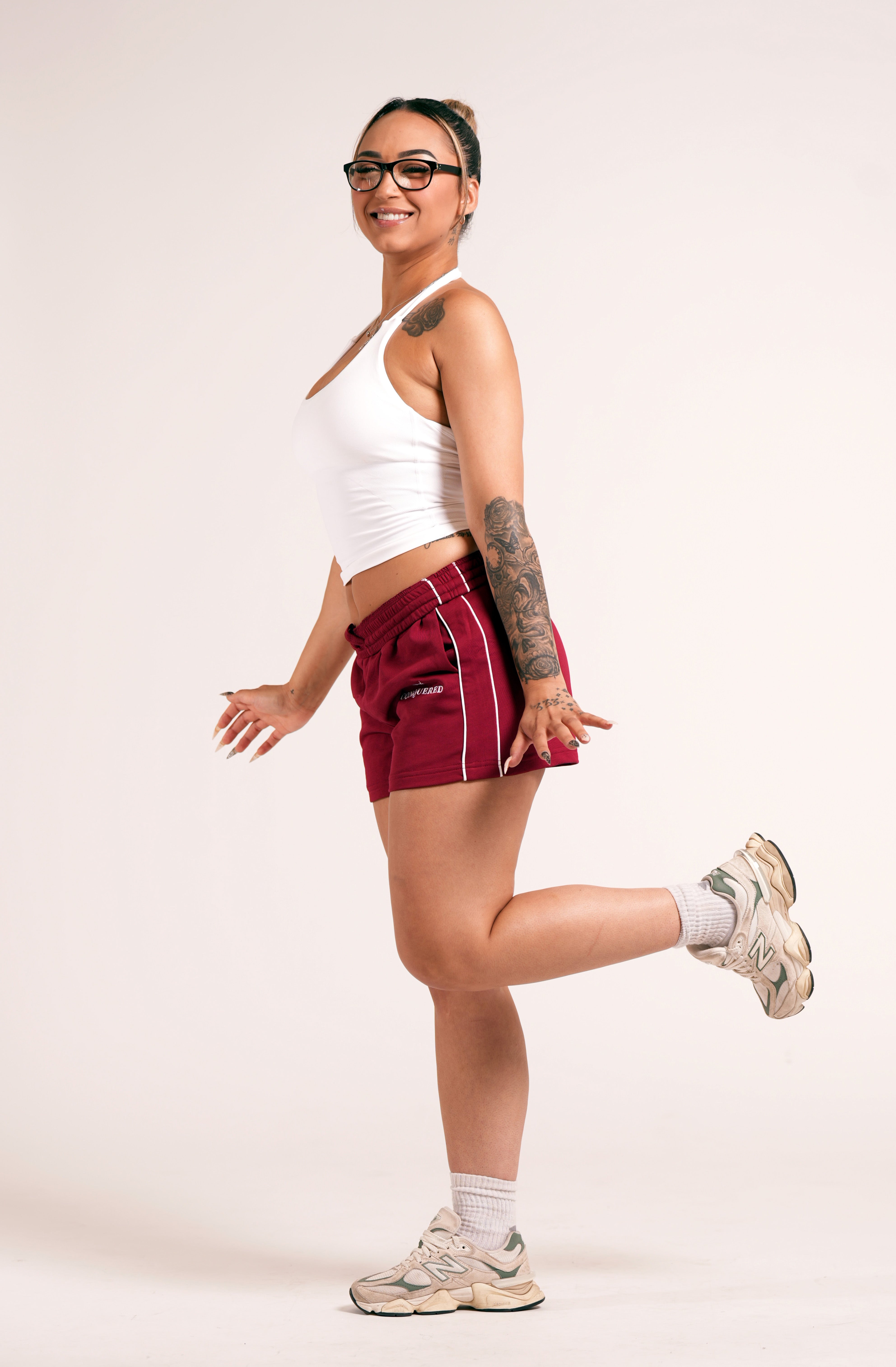 Person wearing crimson red varsity style shorts with white lines on the side with 'Conquered' text and image logo embroidered below the left pocket pairing with a white halter top in front of a plain white background