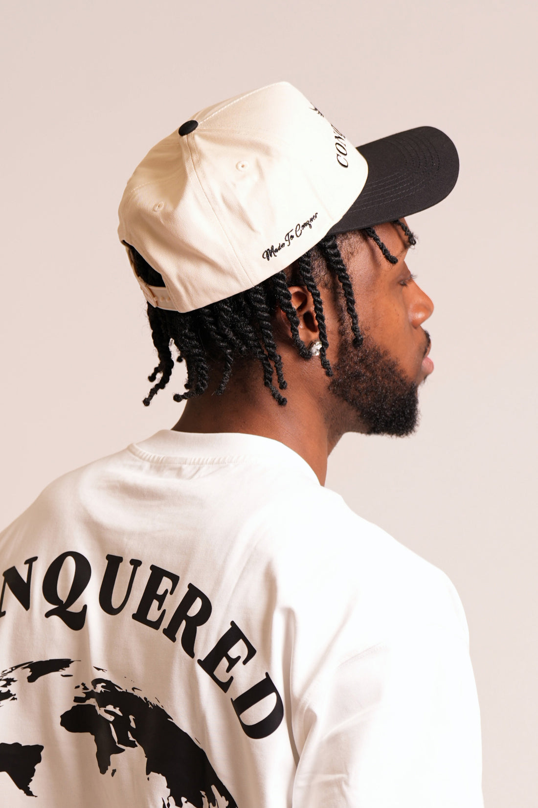 Person wearing a cream and black coloured satin lined cap with 'CONQUERED' word and image branding on a plain white background