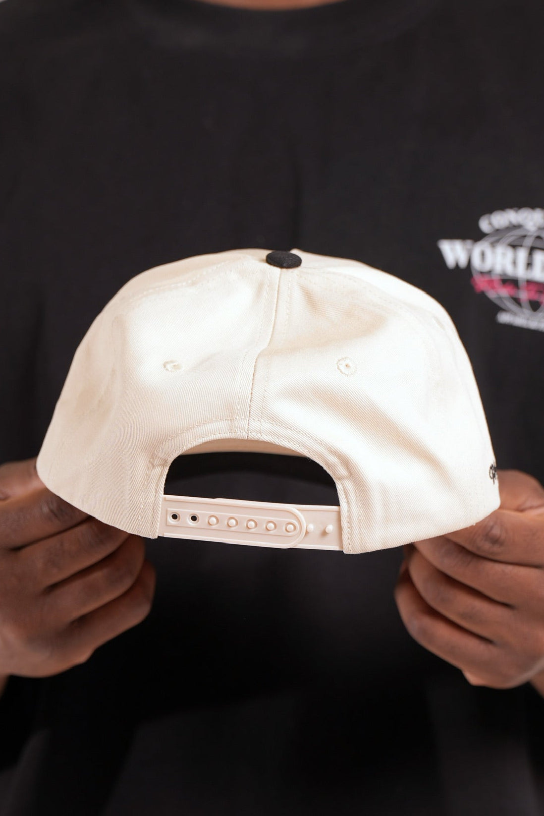 Person holding a cream and black satin lined cap with 'Conquered' text and image logo stitched on the front panel against a black shirt background