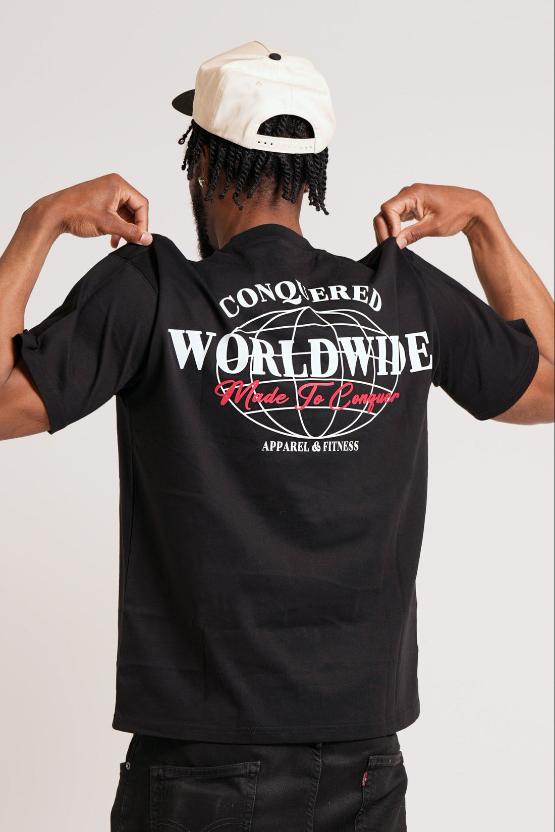 Person wearing a short sleeve black oversized t-shirt with a graphic design that reads "CONQUERED WORLDWIDE" in white and "Made To Conquer" in red on a plain white background