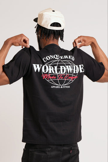 Person wearing a short sleeve black oversized t-shirt with a graphic design that reads "CONQUERED WORLDWIDE" in white and "Made To Conquer" in red on a plain white background