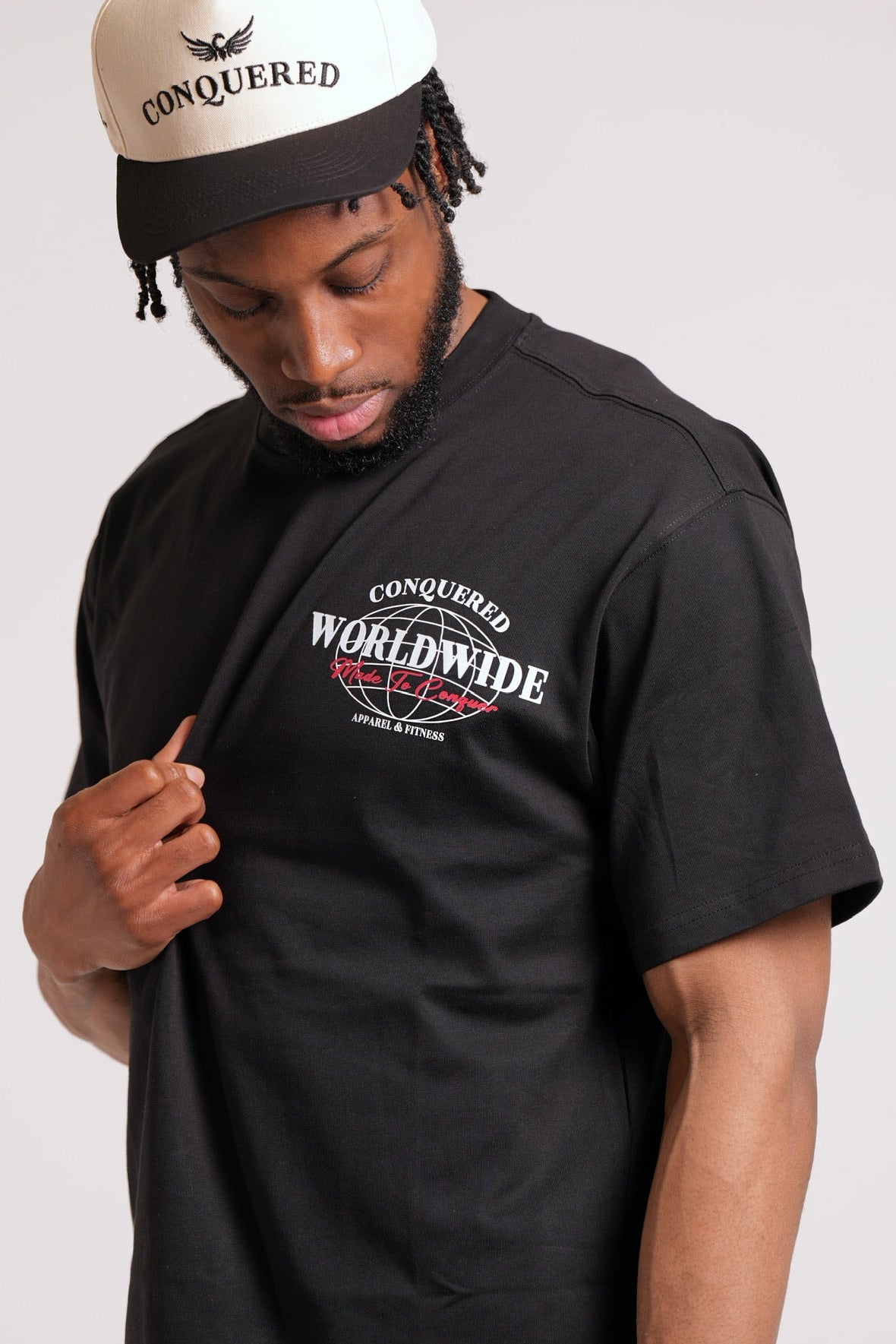 Man wearing a black t-shirt with 'Conquered Worldwide' 'Made To Conquer' graphic and a matching cap on a plain white background