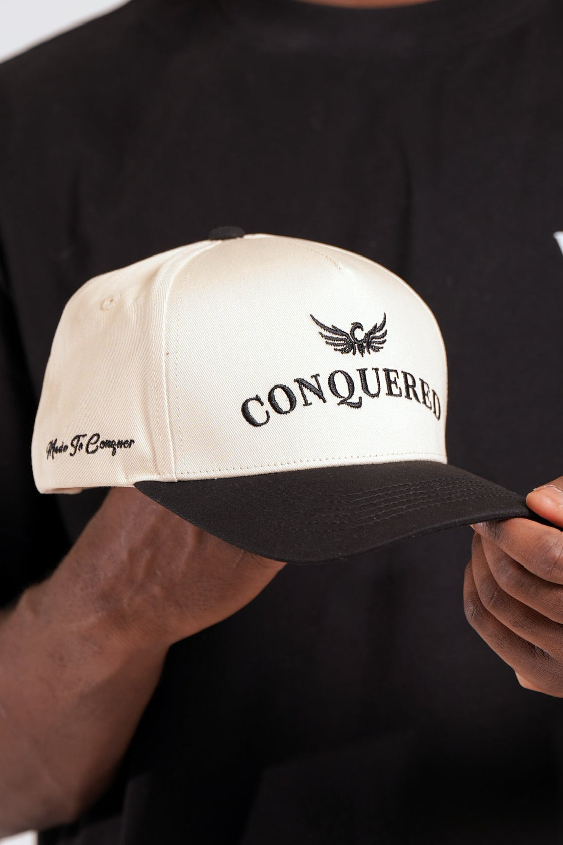 Person holding a cream and black satin lined cap with 'Conquered' text and image logo stitched on the front panel and 'Made To Conquer' embroidered on the right side panel against a black shirt background. 