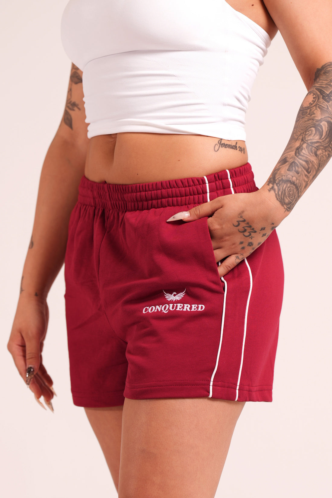 Person wearing crimson red varsity style shorts with white lines on the side with 'Conquered' text and image logo embroidered below the left pocket pairing with a white halter top in front of a plain white background