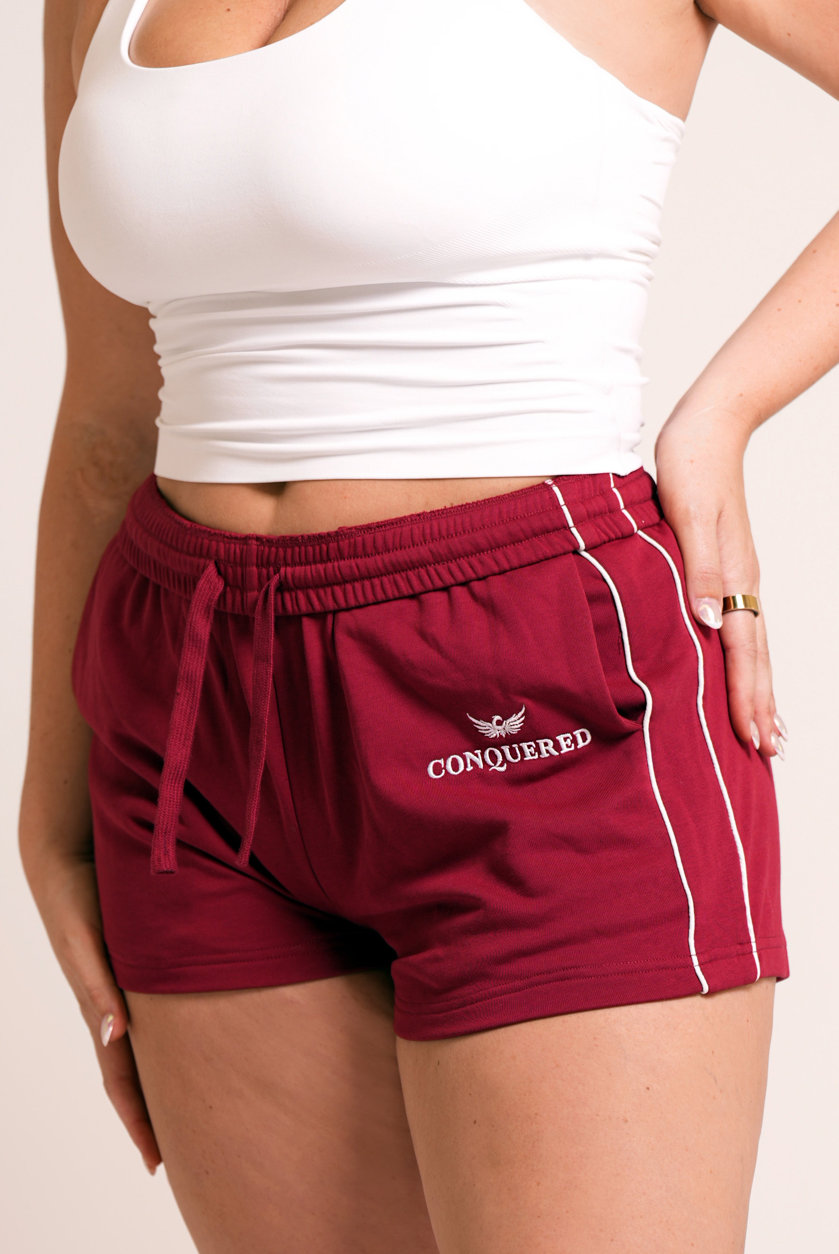 Person wearing crimson red varsity style shorts with white lines on the side with 'Conquered' text and image logo embroidered below the left pocket pairing with a white halter top in front of a plain white background