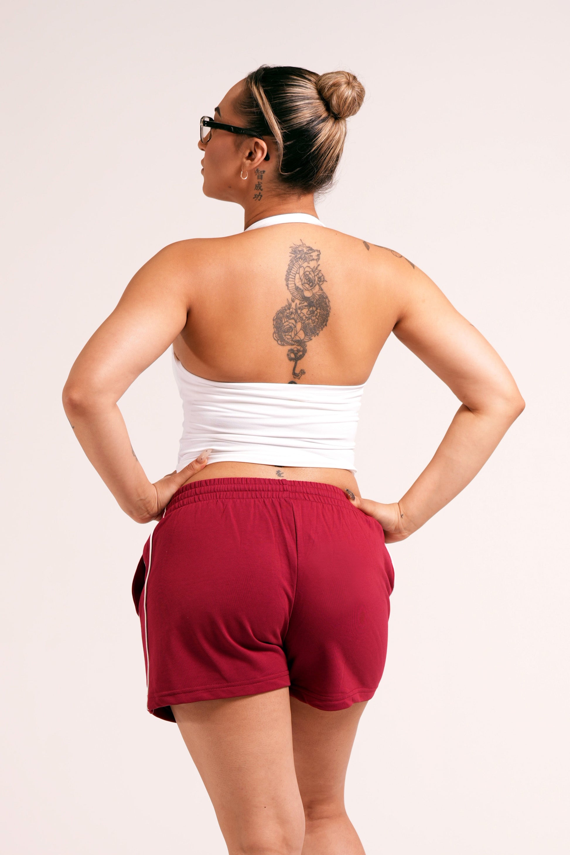 Person wearing crimson red varsity style shorts with white lines on the side with 'Conquered' text and image logo embroidered below the left pocket pairing with a white halter top in front of a plain white background