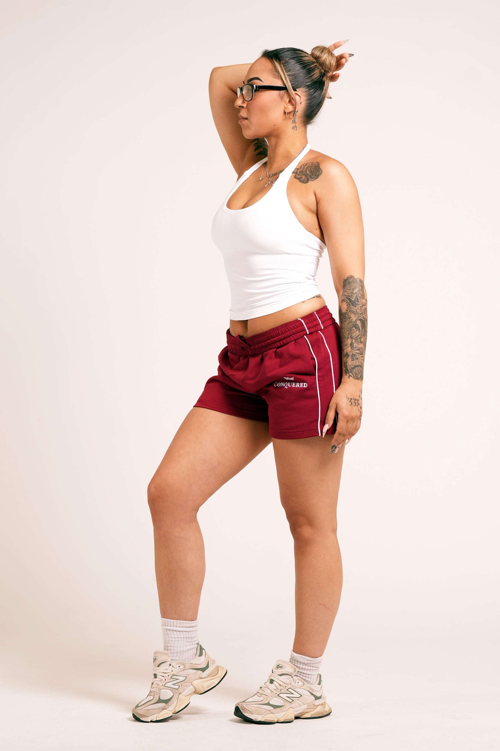 Person wearing crimson red varsity style shorts with white lines on the side with 'Conquered' text and image logo embroidered below the left pocket pairing with a white halter top in front of a plain white background